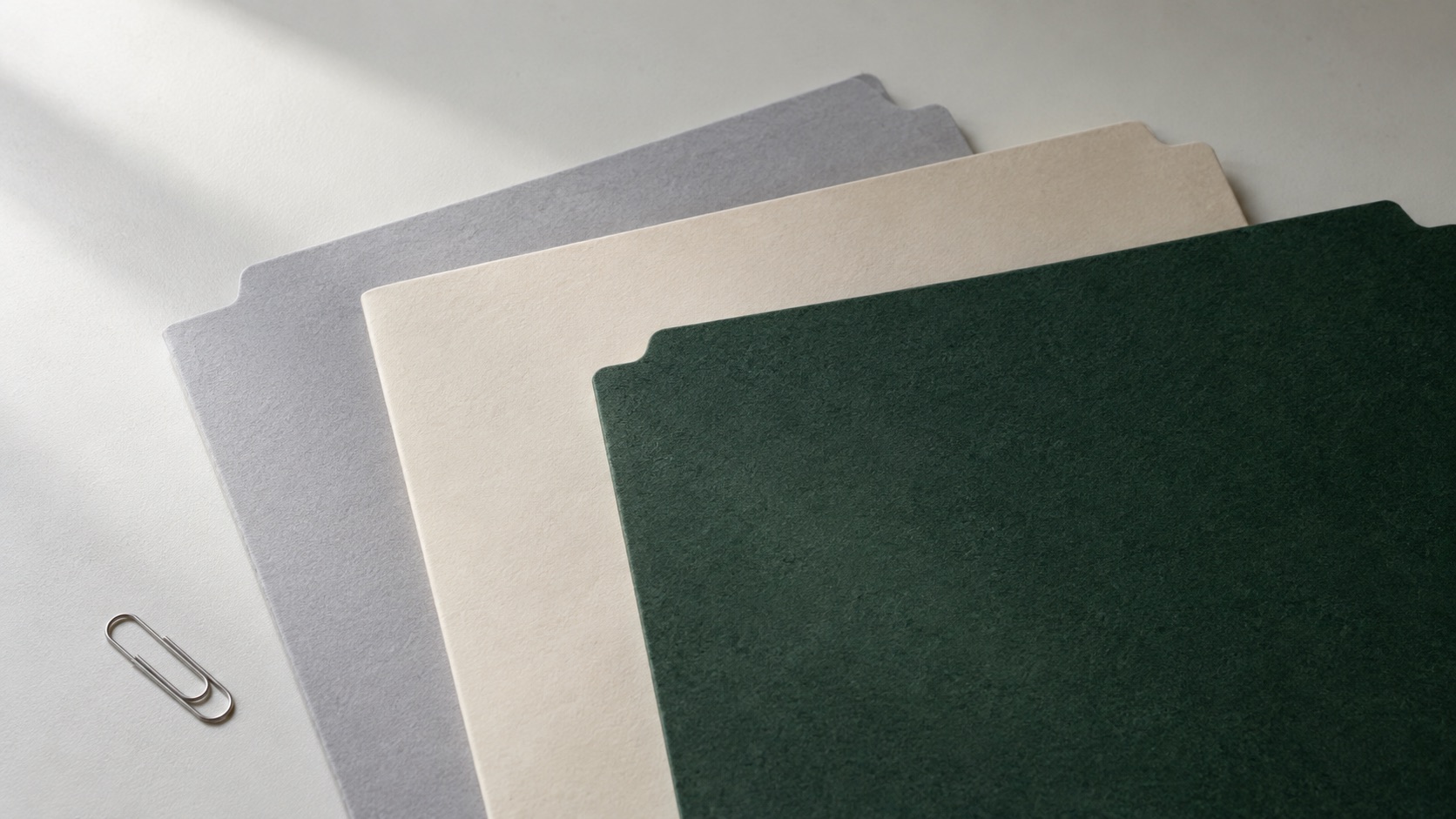 Three partly overlapping plain folders on a desk, with one small metal paperclip